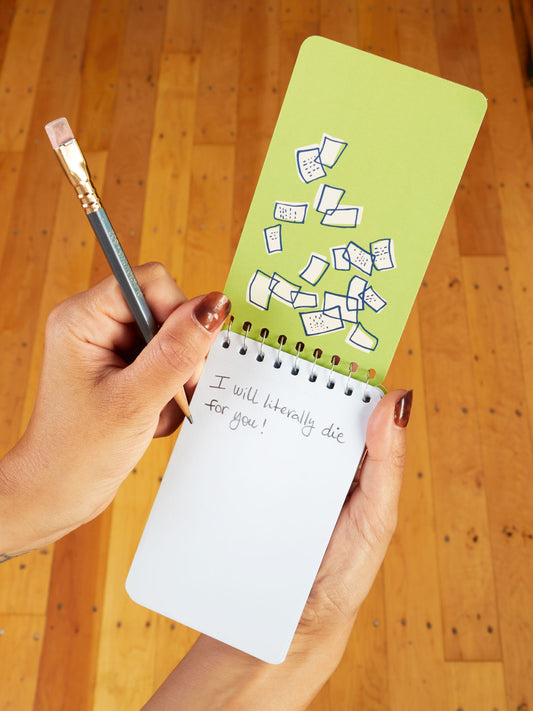 A person, curled in an old blanket, writes "I will literally die for you!" in a Blue Q Deep Thoughts For My Cat To Inherit When I Die 3x5 Spiral Notebook with illustrated cover, deep thoughts drifting over the wooden floor.