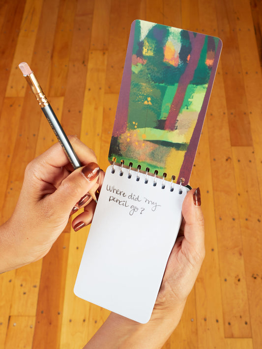 Someone holds a Blue Q "Notes From My Mushroom Trip" 3x5 spiral notebook with an abstract colorful cover and handwritten note, "Where did my pencil go?" next to a pencil eraser on a light wooden floor.