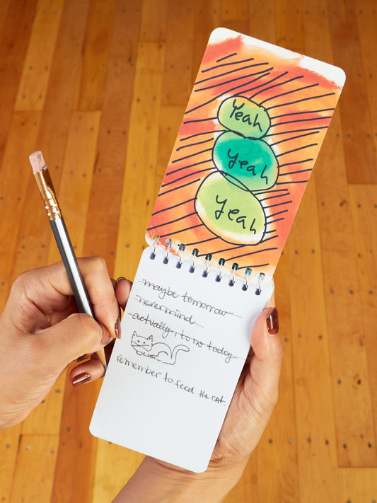 A person holds Blue Q's "The Art Of Procrastination. Have You Seen My Pencil? No, My Good Pencil" 3x5 spiral notebook featuring playful doodles, a witty to-do list, and cat art in a subtle style.