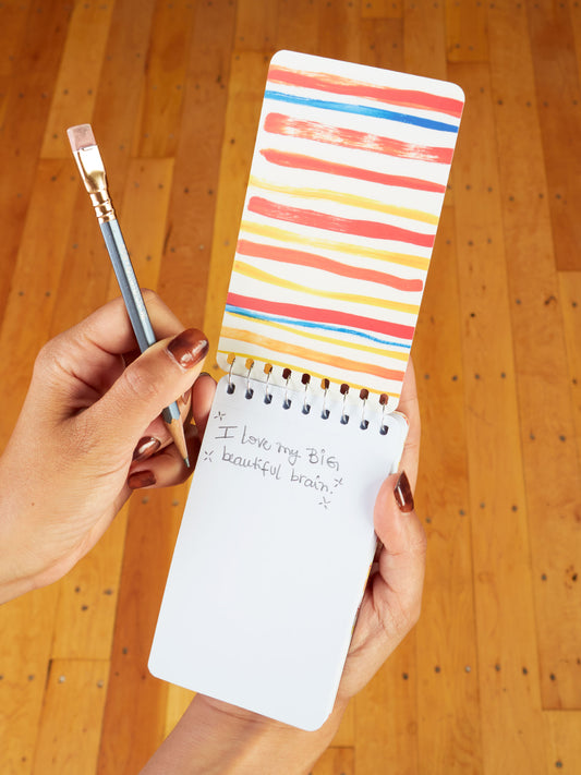 A person with painted nails holds the Blue Q “Preparing For A Busy Day Of Fighting My Mind” 3x5 Spiral Notebook, featuring colorful stripes and handwritten text. They hold a pencil in the other hand, above a wood floor background.