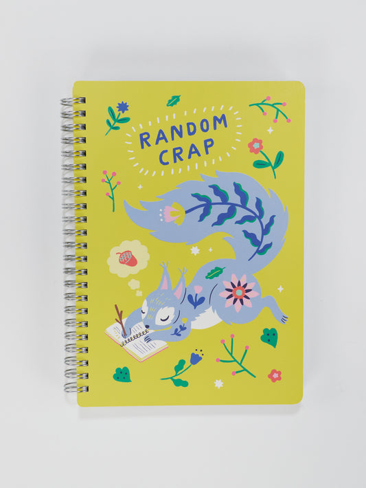 The Blue Q Random Crap 6x8 Spiral Notebook features a yellow cover with a playful blue and purple fox reading among flowers. "RANDOM CRAP" is boldly printed at the top, perfect for jotting down thoughts or storing treasures.