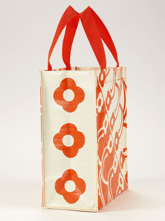 The Blue Q Petal Print Handy Tote is a rectangular bag with red handles, featuring bold red and white floral and geometric designs. This friendly carry tote stands upright on a plain white background.