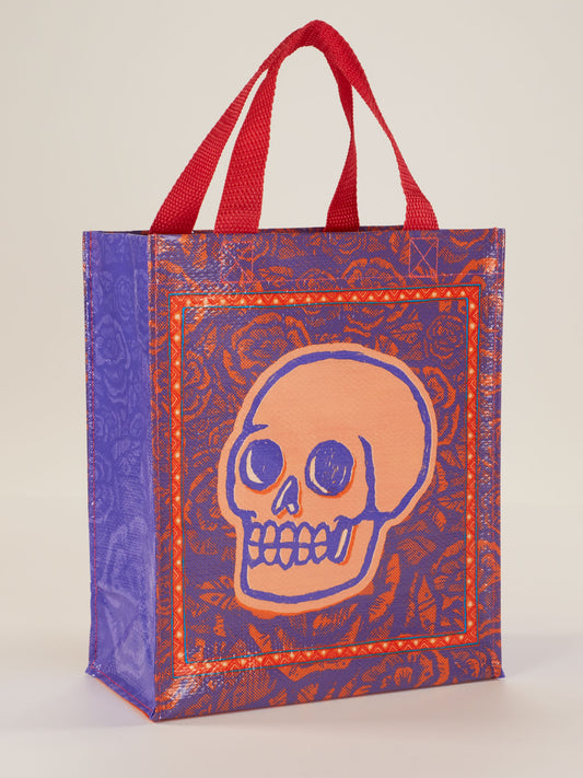 The Skulls & Rose Handy Tote by Blue Q features a striking orange skull with blue outlines on the front, surrounded by a detailed red and purple border. The stylish purple bag comes with bold red handles for both beauty and practicality.