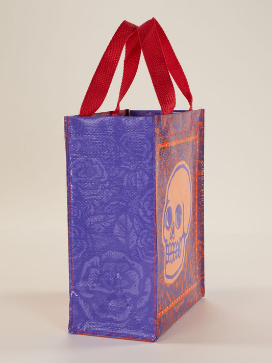 The Blue Q Skulls & Rose Handy Tote is a practical rectangular bag with red handles, a purple background, an orange skull design, and decorative side patterns.