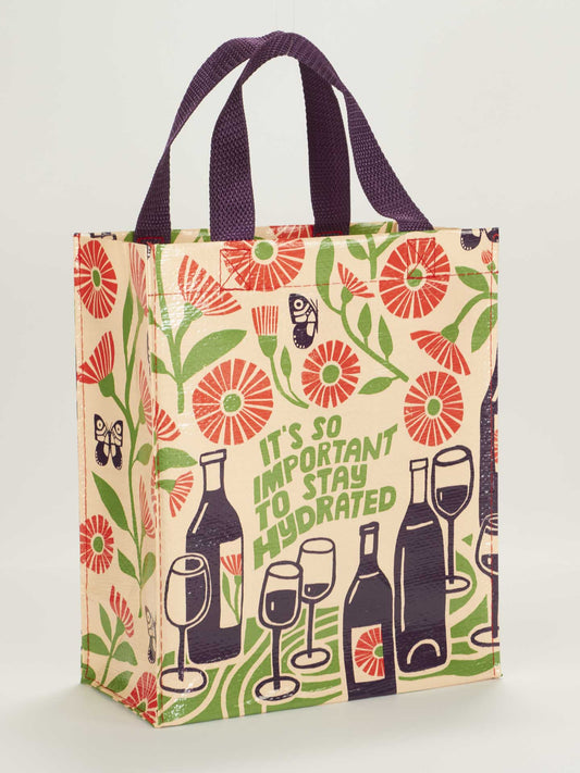 The Blue Q "It's So Important To Stay Hydrated" Handy Tote is a reusable bag with purple handles, wine and floral illustrations, and green lettering of the reminder phrase printed on one side.
