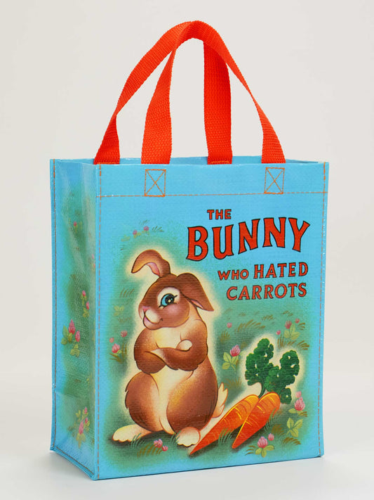 The Bunny Who Hated Carrots Handy Tote by Blue Q is a blue bag with red handles, featuring a bunny illustration with two carrots and green plants, plus the title text for a whimsical storybook vibe.