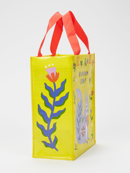 The Blue Q Random Crap Handy Tote is a yellow bag with red handles, floral designs, and a cartoon animal. Made from recycled material, it playfully displays “RANDOM CRAP” on the side—perfect for organizing your stuff.