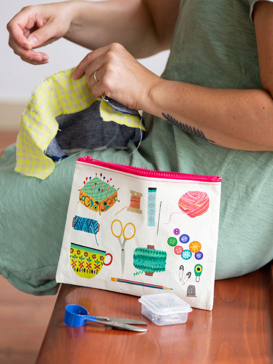 A person in a green dress sews yellow fabric with needle and thread. Nearby, the Blue Q Sewing Kit Zipper Pouch, illustrated with scissors and buttons, sits on a wooden surface surrounded by sewing tools.