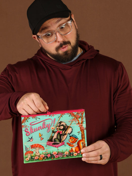 A bearded man in glasses and a black cap, wearing a maroon hoodie, holds up the Skunky Zipper Pouch by Blue Q. Its brown cover features a cartoon skunk on a swing with a squirrel, making for an eye-catching design open to interpretation.