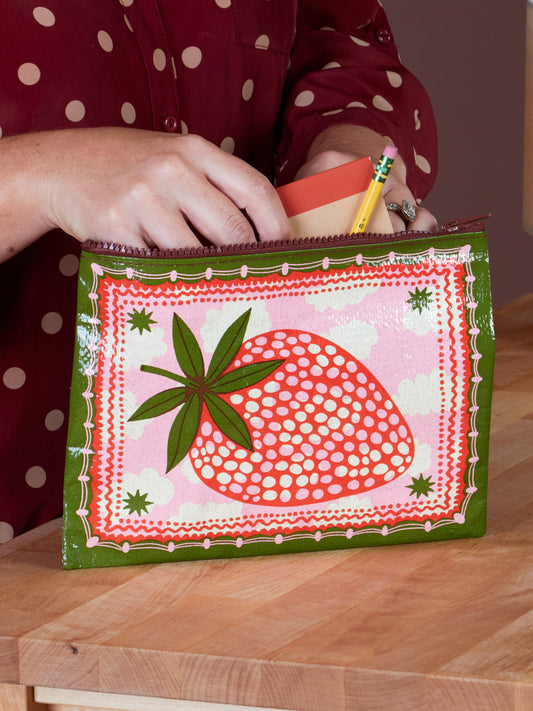 A person puts a pencil and notepad into the Blue Q Strawberry Clouds Zipper Pouch, a colorful case featuring a large red strawberry and green edges reminiscent of ocean grass, set on a wooden surface.