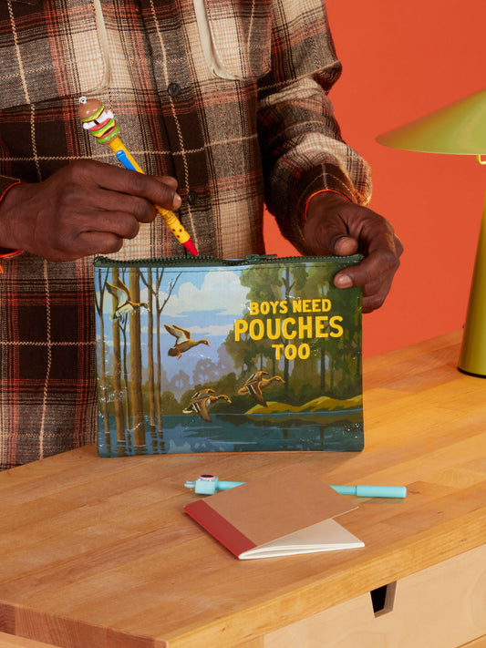 A person in a plaid shirt holds the Blue Q Boys Need Pouches Too Zipper Pouch—featuring ducks and trees—while standing at a wooden table with notebooks, pens, and storage gear, set against an orange background.