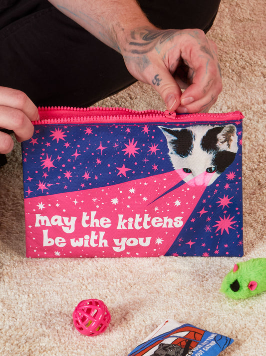 A person unzips the Blue Q "May The Kittens Be With You Zipper Pouch," featuring a cat’s face and playful text on a pink and blue starry design, surrounded by cat toys and a hand-drawn card on a carpeted floor.