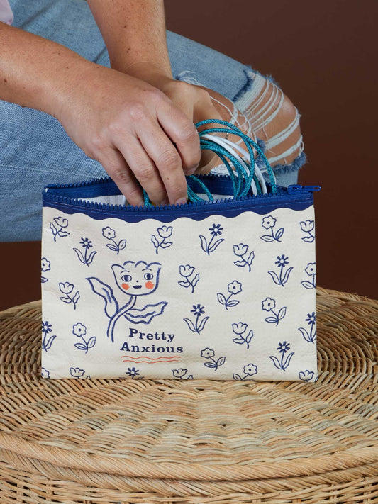 A person puts teal cords into the Blue Q Pretty Anxious Zipper Pouch, which features cream fabric with blue florals, a cat illustration, and the phrase “Pretty Anxious”—proving cute and anxious go together. It sits on a woven surface.