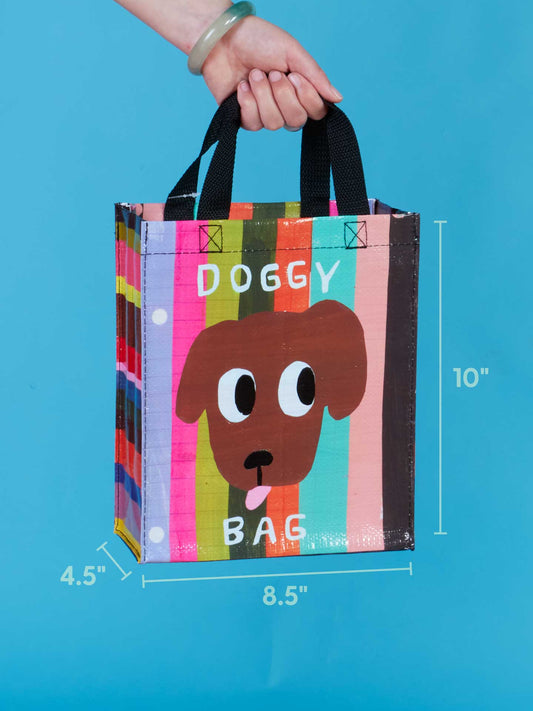 A hand holds the Blue Q Doggy Handy Tote, a cute bag with colorful stripes, black handles, and a cartoon dog face above "DOGGY BAG." Size: 10" tall, 8.5" wide, and 4.5" deep.