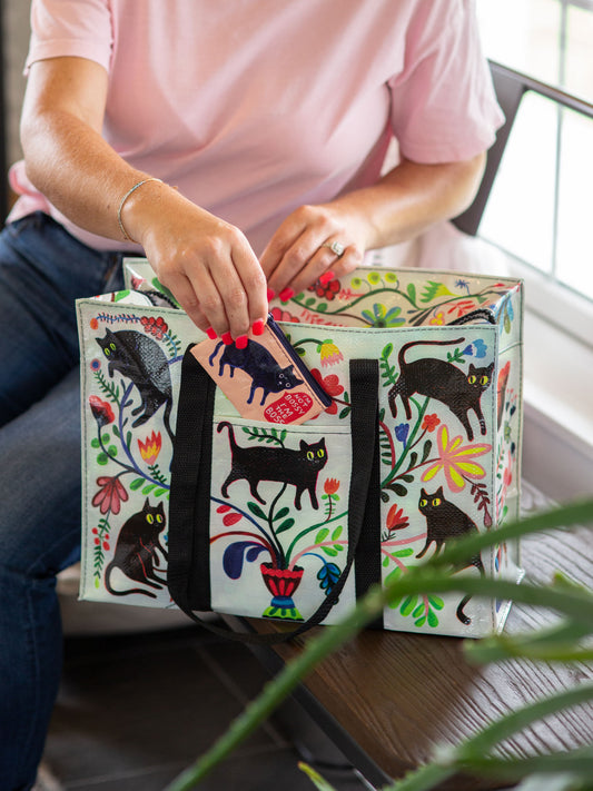 Wearing a pink shirt, someone places the Blue Q "I'm Not Bossy. I'm The Boss" Coin Purse into a vibrant tote adorned with black cats and flowers, showcasing their playful style while seated on a wooden bench.