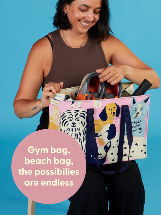 A smiling woman sits on a chair holding the Blue Q Happy Dogs Shoulder Tote, decorated with cheerful dog illustrations and filled with items. A pink circle reads: Gym bag, beach bag—the possibilities are endless.