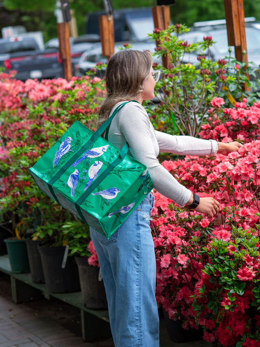A person with long hair, glasses, a white top, and blue jeans stands among blooming pink flowers, carrying the Blue Q Songbird Shoulder Tote over their shoulder.