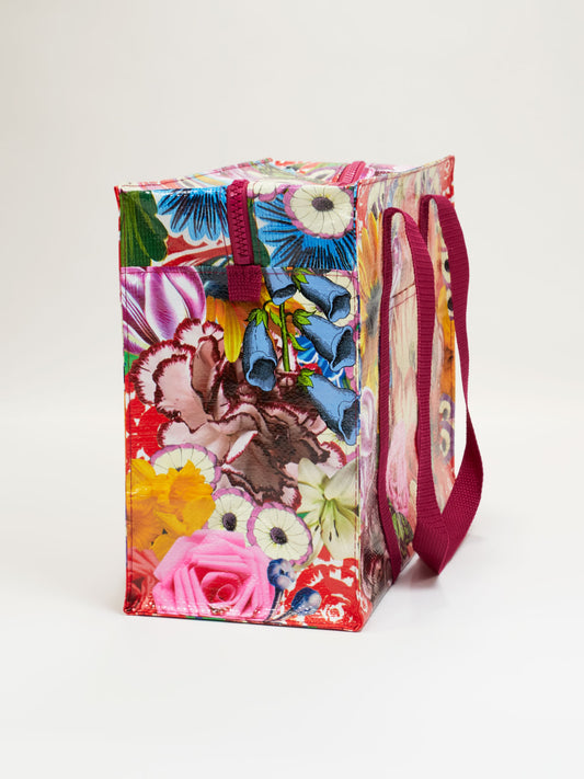 The Blossom Shoulder Tote by Blue Q is a rectangular, reusable bag with a vibrant bouquet-inspired floral print and maroon handles, shown against a plain white background.