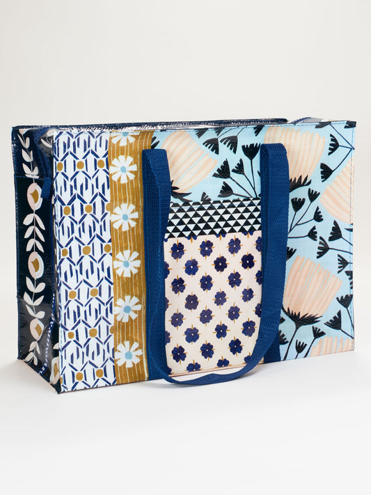 The Patchwork Shoulder Tote by Blue Q is a colorful rectangular bag with strong shoulder straps and blue handles, featuring vibrant floral and geometric patterns in blue, gold, pink, and white.