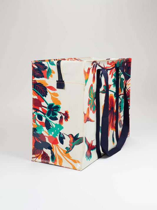 The Blue Q Hummingbird Shoulder Tote is a reusable bag made from recycled material, featuring dark handles and a colorful floral and hummingbird design on a white background.