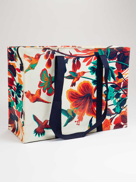 The Blue Q Hummingbird Shoulder Tote is an eco-friendly rectangular bag with dark handles and a vibrant floral and hummingbird design in red, orange, teal, and green on a light background.