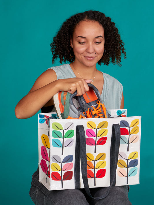 A woman with curly hair smiles as she puts sneakers into the Blue Q Sprout Shoulder Tote, a cute bag featuring colorful leaf patterns and made from recycled materials, set against a teal background.