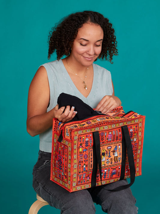 A woman with curly hair smiles as she places a black item into the Blue Q Magic Carpet Shoulder Tote, a colorful patterned recycled bag with black handles, while sitting on a stool against a teal background.