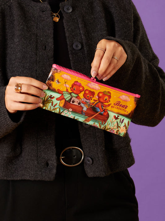 A person in dark clothing holds the Blue Q Bear Necessities Pencil Case, a colorful zipper pouch decorated with cartoon bears and comic book-inspired designs, while sitting in a boat on a flower-filled pond.