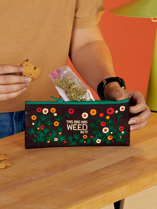 A person holds a cookie and dried herbs beside a Blue Q "This Bag Has Weed In It" Pencil Case, featuring colorful flowers, on a wooden table with a green lamp in the background.