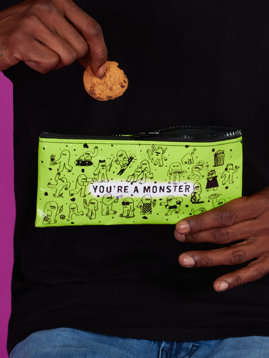 A person holds a cookie above the Blue Q You're A Monster Pencil Case, a green pouch with monster doodles and "MY monster" text. They're wearing a black shirt and blue jeans.