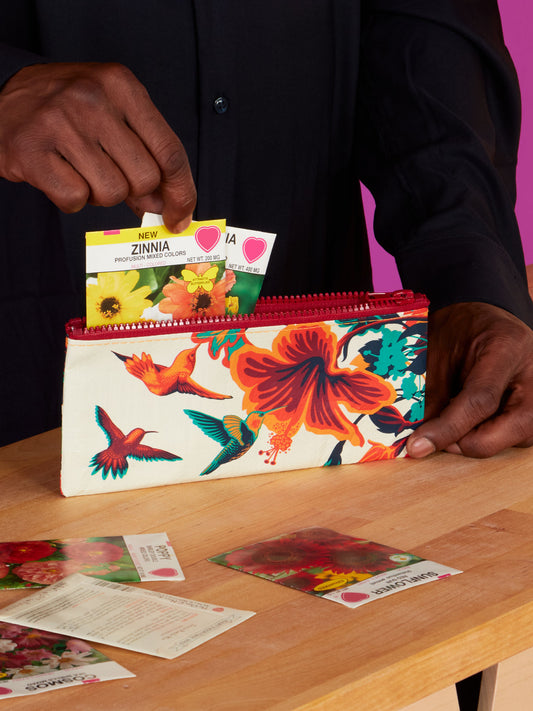 A person puts a Blue Q Hummingbird Pencil Case, featuring bright colors, on a wooden surface with other items nearby.