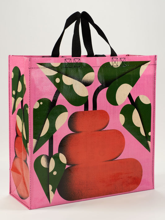 The Blue Q Bubble Vase Shopper is a reusable bag with black handles, decorated with curvy red stone-like shapes and green leafy accents with white spots on a vibrant pink background.