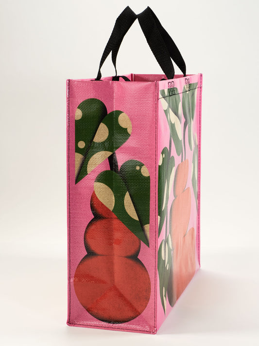 The Bubble Vase Shopper by Blue Q is a reusable shopping bag with black handles, showcasing a pink background and vibrant curvy green leaves and red shapes on the sides.