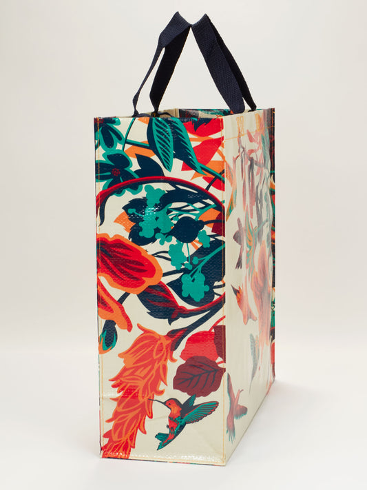 The Blue Q Hummingbird Shopper is a reusable shopping bag with dark blue handles, featuring a colorful botanical pattern with leaves, flowers, and hummingbirds on a white background—ideal for stylish everyday use.