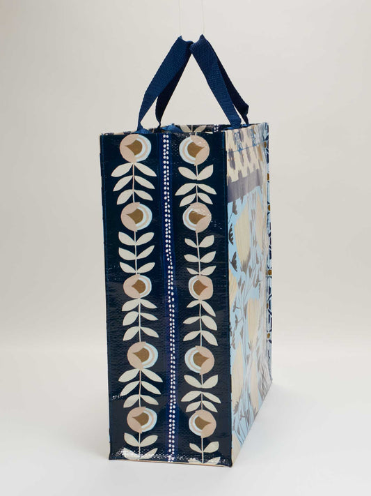The Blue Q Patchwork Shopper is a rectangular, spacious bag with navy blue sides, dark blue handles, and a light floral patterned front featuring beige and white stylized flowers and leaves in vertical rows. Reusable for storage or shopping.