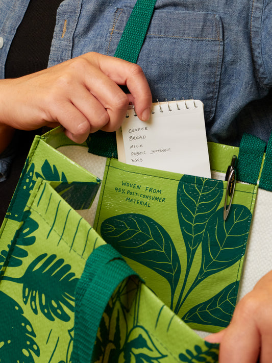 A person places a small notepad with a grocery list into the Blue Q "I Have A Bit Of A Plant Problem Shopper," a green reusable bag with leaf patterns, made from 95% post-consumer material.