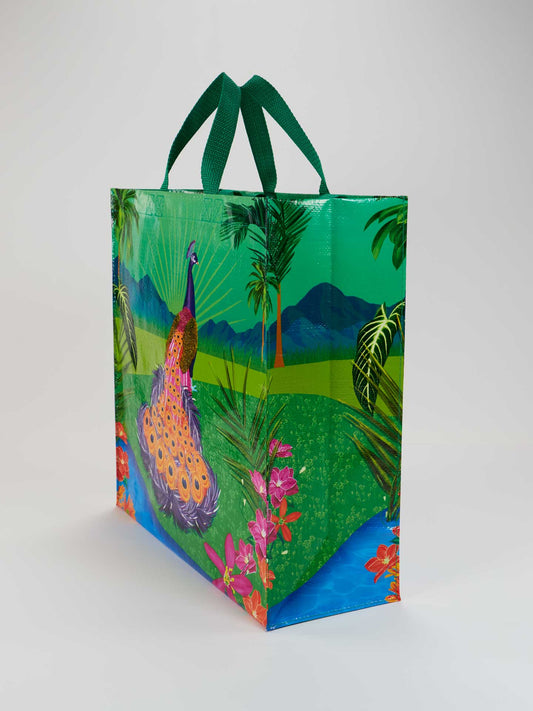 The Peacock Paradise Shopper by Blue Q is a reusable tote bag made from recycled material, featuring green handles and a vibrant peacock among flowers, palm trees, and mountains under a blue sky.