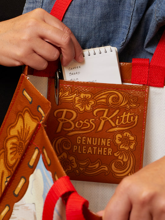 A person slips a small notepad with a grocery list into the pocket of a Boss Kitty Shopper by Blue Q, a brown reusable bag featuring red straps, decorative floral patterns, and “Boss Kitty Genuine Pleather” embossed on the front.