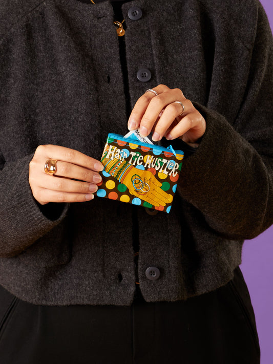 A person in a black coat holds the Blue Q Hair Tie Hustler Coin Purse, featuring polka dots and a cartoon hand design. "HAIR TIE HUSTLER" is printed on it. The purple background makes its vibrant colors stand out.
