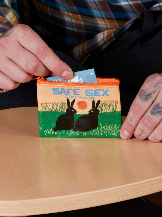 A person places a condom packet into the Blue Q Safe Sex Coin Purse, featuring an image of two black rabbits in grass beneath an orange sun.