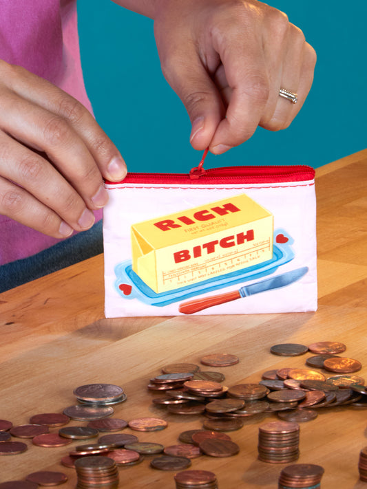 A person zips the Blue Q Rich Bitch Coin Purse, featuring a butter stick illustration with "RICH BITCH" text, above scattered coins on a wooden table—an ideal quirky gift for fun-loving friends.