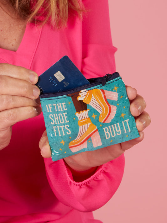 A person in a pink top holds the Blue Q "If The Shoe Fits, Buy It Coin Purse," featuring retro roller skates and the phrase, while placing a blue credit card inside.