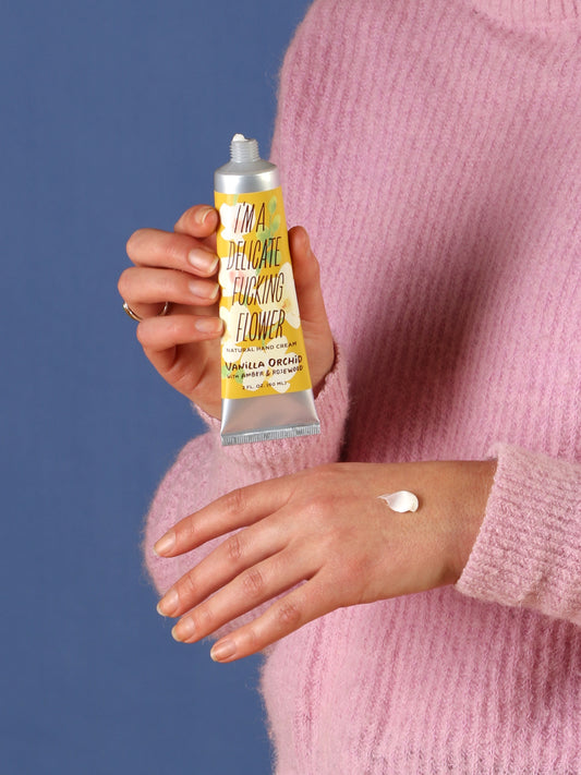 A person in a pink sweater squeezes Blue Q's I'm A Delicate Fucking Flower Hand Cream—Vanilla Orchid with Amber Rosewood—from its tube onto their hand, set against a blue background.