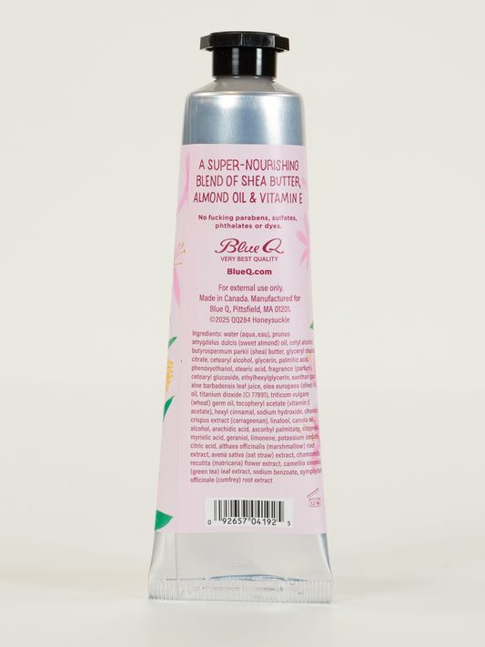 A pink and silver tube of Blue Q’s “I'm a Delicate Fucking Flower” Honeysuckle Hand Cream stands upright, showing the back label with shea butter, almond oil, vitamin E, barcode, and details about nourishing marigold & wild freesia.