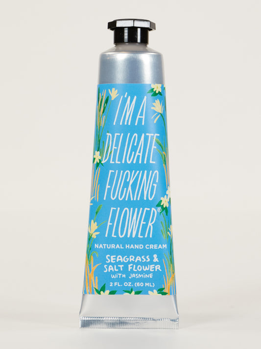 A blue tube of Blue Q “I'm a Delicate Fucking Flower” Natural Hand Cream–Sea Grass & Salt Flower with jasmine, floral design, shea butter, and the label “2 FL. OZ. (60 mL).”.