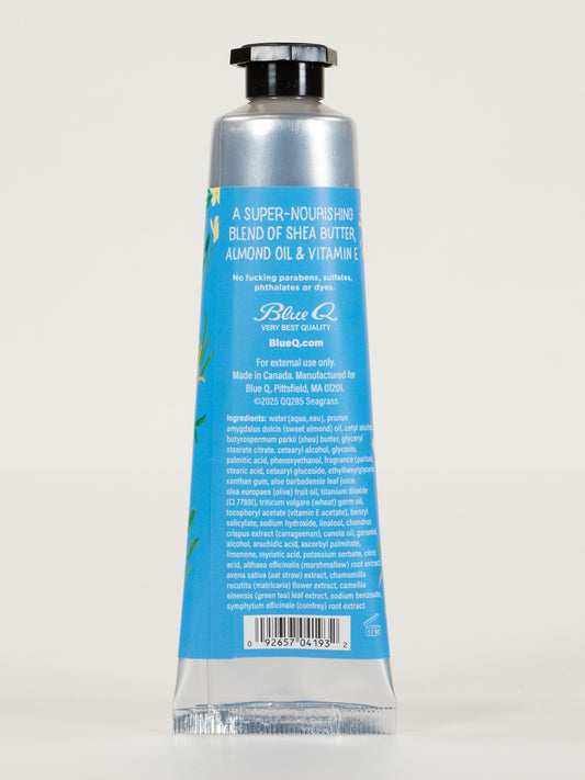The image shows the back of a Blue Q "I'm a Delicate Fucking Flower" hand cream tube in silver and blue with a black cap, listing ingredients like shea butter, almond oil, vitamin E, product details, and barcode. Scent: Sea Grass & Salt Flower With Jasmine.