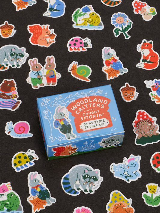 A blue box labeled Blue Q’s Woodland Critters Caught Smokin’ Playtime Sticket Set rests surrounded by charming critters—colorful animal stickers featuring raccoons, deer, snails, and rabbits—set against a black background. Perfect for ages 13 and up, this delightful set is ideal for fans of whimsical designs and collectible figurine-inspired style.