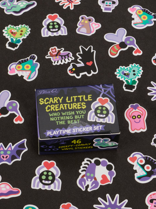 A box labeled Blue Q’s Scary Little Creatures Who Wish You Nothing But The Best Playtime Sticker Set sits on a surface scattered with colorful, spooky cartoon creature stickers. Designed for ages 13 and up, the box promises 46 creepy-crawly vinyl stickers inside—perfect for anyone who believes even misunderstood monsters deserve a playful moment.