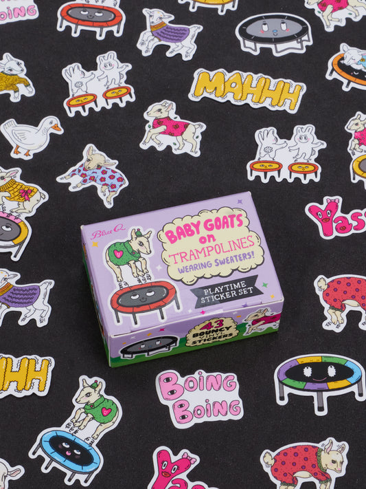 A box labeled “Baby Goats On Trampolines Wearing Sweaters Playtime Sticker Set” by Blue Q rests on a black surface, surrounded by colorful stickers featuring goats on trampolines, geese, playful sound effect words, and more—a perfect novelty gift for ages 13 and up.