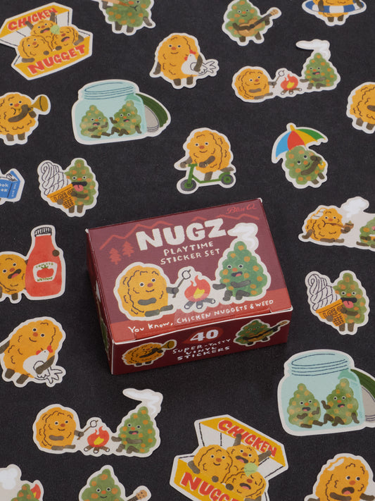 A box labeled Nugz Playtime Sticker Set by Blue Q rests on a black surface scattered with vibrant stickers featuring chicken nuggets, cannabis buds, ketchup, and whimsical cartoon scenes—ideal accessories for teens who want to add a playful touch to their dabber and dipper collections.